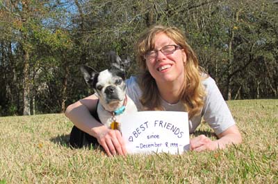 Amanda Gene lies on the ground with her arm wrapped around Noodles. Noodles is a small black and white Boston Terrier. She holds a sign that says "Best Friends."